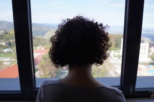 A woman with curly hair gazing out a window at a scenic mountain view, indoors.