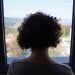 A woman with curly hair gazing out a window at a scenic mountain view, indoors.